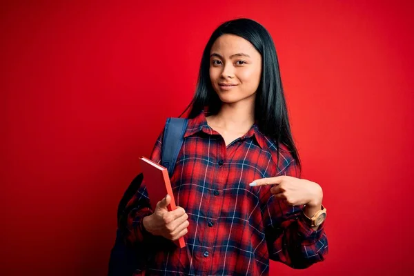 年轻美丽的中国女学生手拿着书本站在孤立的粉色背景上 非常高兴地用手和手指指指点点 — 图库照片