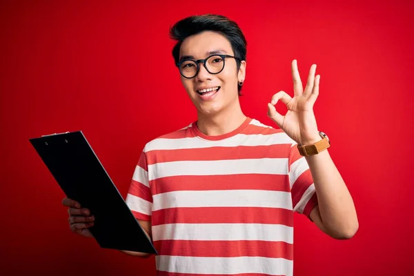 Young handsome chinese inspector man wearing glasses holding clipboard checklist doing ok sign with fingers, excellent symbol