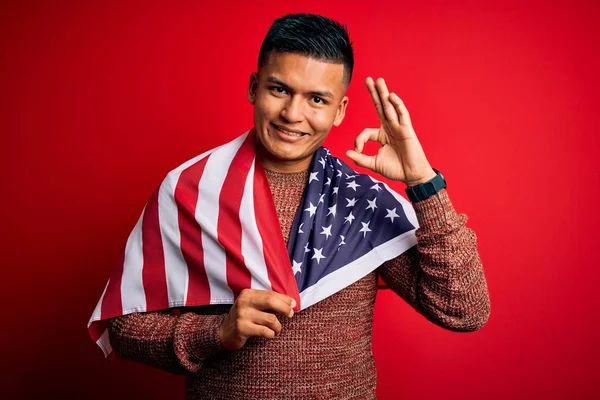 Young handsome latin patriotic man wearing united states of america falg doing ok sign with fingers, excellent symbol