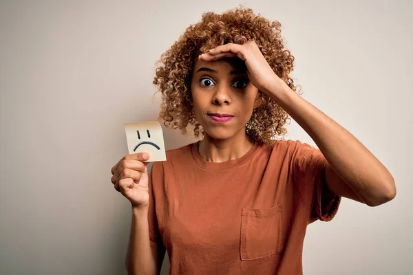 Young african american woman with curly hair holding reminder paper with sad face emoji stressed with hand on head, shocked with shame and surprise face, angry and frustrated. Fear and upset for mistake.