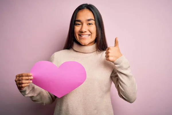 Young beautiful asian woman holding pink heart standing over isolated background happy with big smile doing ok sign, thumb up with fingers, excellent sign