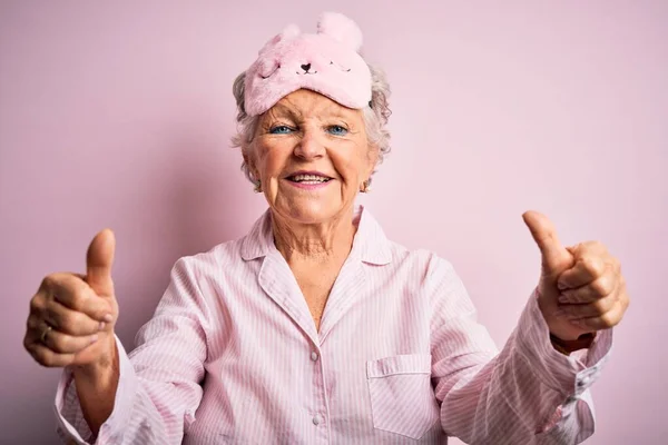 上了年纪的漂亮女人 戴着睡眠面罩 披着睡衣 有着淡淡的粉色背景 她赞许地用手做着积极的手势 微笑着竖起大拇指 为成功感到高兴 赢的手势 — 图库照片