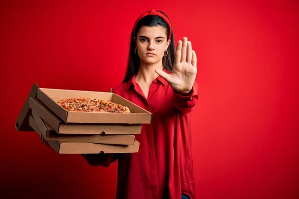 若いです美しいですブルネット女性を保持していますデリバリーボックス上イタリアのピザとともに赤の背景にオープン手を行います停止標識で真剣で自信を持って表現 防衛ジェスチャー — ストック写真