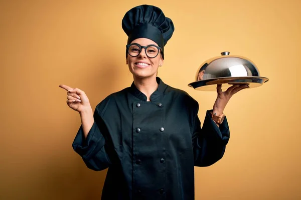 Young beautiful brunette chefwoman wearing cooker uniform and hat holding tray with dome very happy pointing with hand and finger to the side