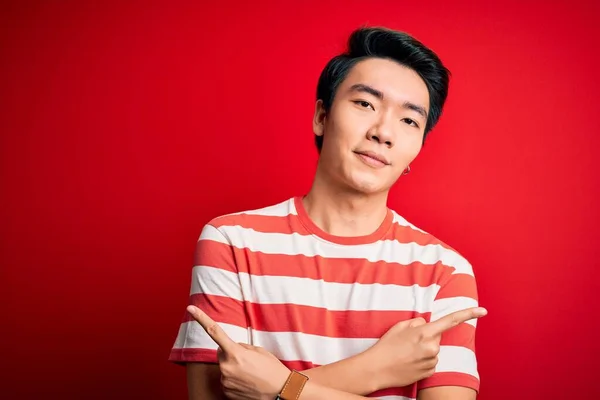 若いですハンサムな中国人男性を着てカジュアルな縞模様のTシャツ赤い背景の上に立って指で両側を指して 異なる方向が反対 — ストック写真