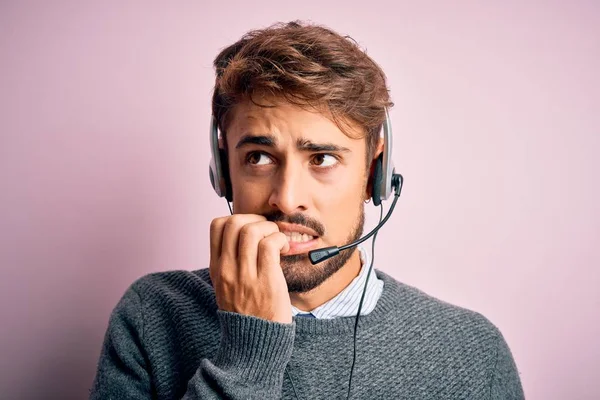 Young call center agent man with beard wearing headset over isolated pink background looking stressed and nervous with hands on mouth biting nails. Anxiety problem.