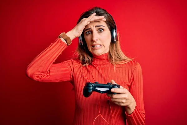 Young beautiful brunette gamer woman playing video game using joystic and headphones stressed with hand on head, shocked with shame and surprise face, angry and frustrated. Fear and upset for mistake.