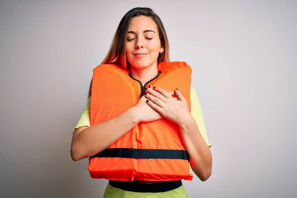 Young beautiful blonde woman with blue eyes wearing orange lifejacket over white background smiling with hands on chest with closed eyes and grateful gesture on face. Health concept.