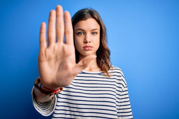 Young beautiful blonde girl wearing casual sweater standing over blue isolated background doing stop sing with palm of the hand. Warning expression with negative and serious gesture on the face.