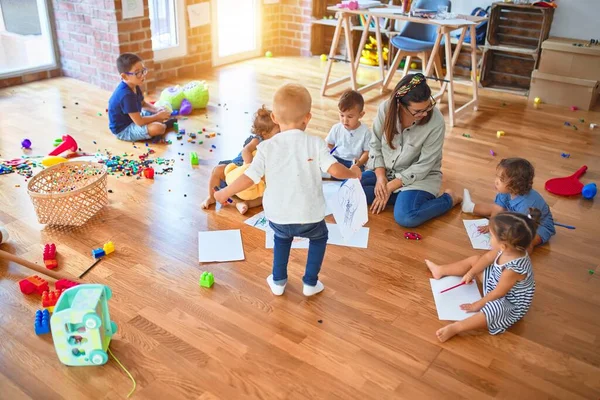 Beautiful teacher and group of toddlers sitting on the floor drawing using paper and pencil around lots of toys at kindergarten