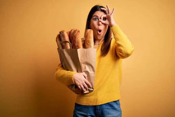 年轻美丽的女人拿着一袋黄色背景的新鲜健康的面包 做着一个很好的手势 脸上带着惊讶的表情 用手指看着 难以置信的表情 — 图库照片