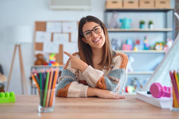 Young beautiful teacher woman wearing sweater and glasses sitting on desk at kindergarten cheerful with a smile on face pointing with hand and finger up to the side with happy and natural expression