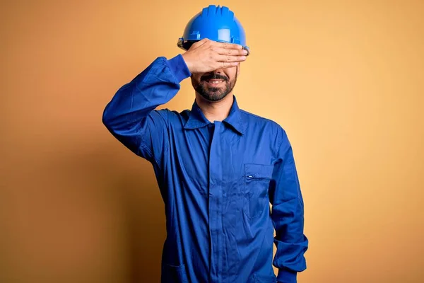 Mechanic man with beard wearing blue uniform and safety helmet over yellow background smiling and laughing with hand on face covering eyes for surprise. Blind concept.