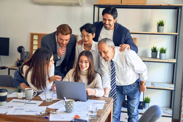 Group of business workers smiling happy and confident. One of them sitting and partners standing around. Working together with smile on face looking at the laptop at the office