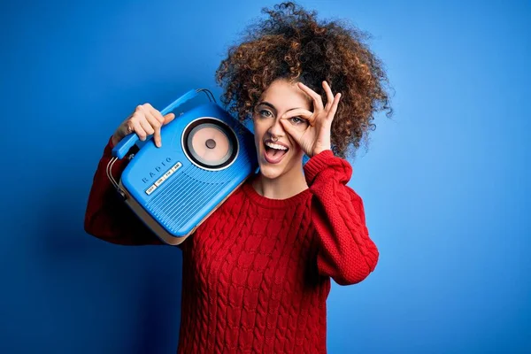 Young beautiful woman with curly hair and piercing listening to music using vintage radio with happy face smiling doing ok sign with hand on eye looking through fingers