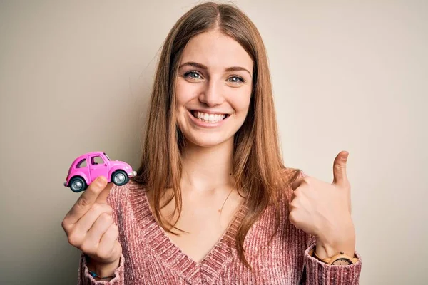 若い美しい赤毛の女性は大きな笑顔で幸せな孤立した白い背景の上に小さなピンクの車を保持します — ストック写真