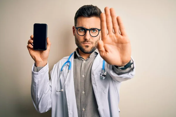 年轻医生 手持听诊器 在孤立的背景下展示智能手机屏幕 张开手 做停止手势 表情严肃自信 防守姿势 — 图库照片