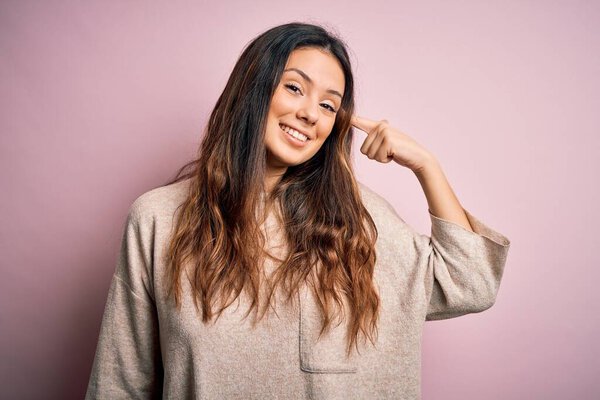 Молодая красивая брюнетка женщина в обычном свитере стоя на розовом фоне улыбаясь указывая на голову одним пальцем, отличная идея или мысль, хорошая память
