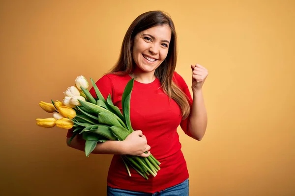 Young blonde woman holding romantic bouquet of tulips flowers over yellow background very happy and excited doing winner gesture with arms raised, smiling and screaming for success. Celebration concept. — Stock Image Young Blonde Woman Holding Romantic Bouquet Tulips Flowers Yellow Background Royalty Free Stock Photos