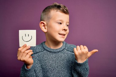Küçük beyaz bir çocuk gülen suratını kâğıt bir notta göstererek mutlu bir mesaj veriyor ve gülümseyen bir yüzle baş parmağıyla işaret ediyor.