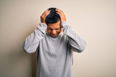 Genç, yakışıklı, latin kazaklı bir adam izole edilmiş beyaz arka planda dikiliyor. Ağrı ve migren yüzünden baş ağrısı ve stres altında. Ellerini başının üstüne koy..