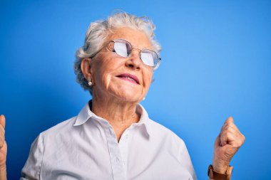 Üst düzey, zarif gömlek ve gözlük takan güzel bir kadın izole edilmiş mavi arka planda çok mutlu ve heyecanlı bir şekilde kollarını kaldırıp, gülümseyerek ve başarı için çığlık atarak kazanan bir jest yapıyor. Kutlama konsepti.