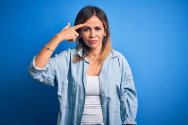 Orta yaşlı, rahat gömlekler giyen, mavi arka planda duran güzel bir kadın alnındaki sivilceden mutsuz olduğunu gösteriyor, siyah noktalı çirkin bir enfeksiyon. Sivilce ve deri problemi