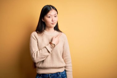 Sarıya boyanmış arka planda sıradan bir süveter giyen genç, güzel Asyalı bir kadın. El parmağı yanda, ciddi ve sakin bir yüz ifadesi sergiliyor.