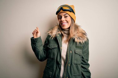 Genç esmer kayakçı kadın kar giysileri ve kayak gözlükleriyle beyaz arka planda, yüzünde kocaman bir gülümsemeyle kameraya el ile işaret ediyor..