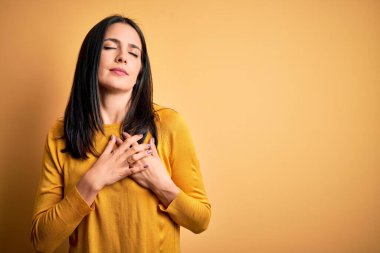 Mavi gözlü genç esmer kadın sarı arka planda rahat bir süveter giyiyor elleri göğsünde, gözleri kapalı ve yüzünde minnet dolu bir jest ile gülümsüyor. Sağlık kavramı.