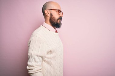 Sakallı, dövmeli, pembe arka planda gözlüklü ve süveterli yakışıklı, rahat bir görünüm takınan, kendine güvenen bir gülümsemesi olan kel bir adam..