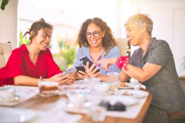 Orta yaşlı kadınlarla buluşup, öğle yemeği yiyip, kahve içiyorlar. Olgun arkadaşlar mutlu bir şekilde gülümsüyor. Güneşli bir günde evde akıllı telefon kullanıyorlar.