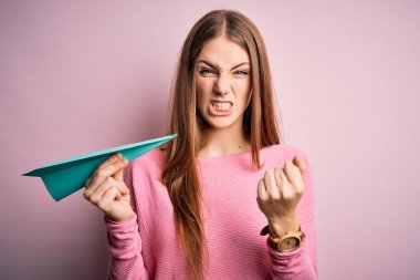 Genç, güzel, kızıl saçlı bir kadın pembe arka planda kâğıt uçak tutuyor öfkeyle bağırıyor, çıldırıyor ve öfkeyle bağırıyor.
