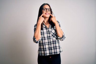 Mavi gözlü genç esmer kadın beyaz arka planda günlük gömlek ve gözlük takıyor ağzı açık gülümsüyor, parmakları işaret ediyor ve zorla neşeli bir gülümseme sergiliyor.