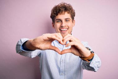 Sarışın, kıvırcık saçlı, beyaz arka planda çizgili tişört giyen, elleri ile kalp sembolü yapan genç, sarışın bir adam. Romantik konsept.