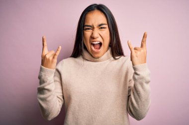 Genç ve güzel Asyalı kadın pembe arka plan üzerine balıkçı yaka kazak giyip çılgın bir ifadeyle elleri havada rock sembolü yaparak bağırıyor. Müzik yıldızı. Ağır konsept.