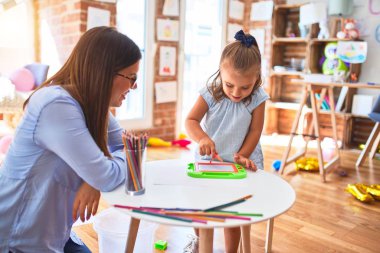 Beyaz kız çocuk, bayan öğretmenle oyun oynayıp öğreniyor. Anne ve kızı oyun odasında oyuncakların etrafında manyetik karatahtaya resim çiziyorlar.