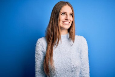 Genç, güzel, kızıl saçlı bir kadın izole edilmiş mavi arka plan üzerine günlük süveter giyip yüzünde gülümseme, doğal bir ifade ile yan yana bakıyor. Kendinden emin gülen.