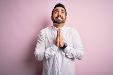 Sakallı genç yakışıklı bir adam pembe arka planda dikilip dua ediyor ve yalvarıyor. Yüzünde umut ifadesi var. Çok duygusal ve endişeli. Yalvarıyorum..