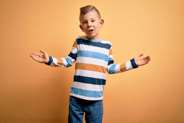 Küçük, beyaz, mavi gözlü, sarı arkaplan üzerine renkli çizgili gömlek giyen kolları ve elleri havada, kafası karışmış bir çocuk. Şüphe kavramı.