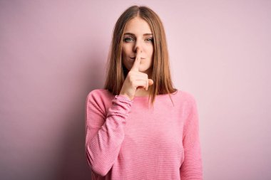 Genç, güzel, kızıl saçlı bir kadın pembe arka planda rahat bir süveter giyiyor ve dudakları parmaklarıyla sessiz olmak istiyor. Sessizlik ve gizli konsept.