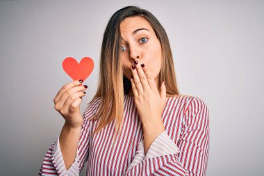 Genç, sarışın, romantik bir kadın, beyaz arka planda kırmızı kağıt kalp taşıyor. Ağzı kapalı, utancından ağzı açık, korkusunun dışa vurumu, sessizlikten korkması, gizli konsept.
