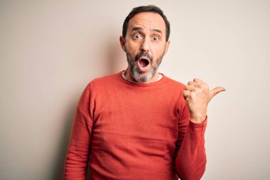 Orta yaşlı, turuncu süveter giyen, izole edilmiş beyaz arka planda dikilen, el parmağı yanda duran, ağzı açık, şaşırmış bir adam..