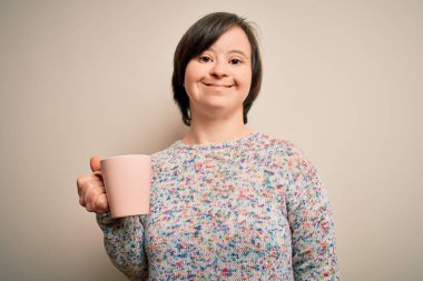 Genç Down sendromlu kadın izole edilmiş arka planda bir fincan sıcak kahve içiyor. Mutlu bir yüzü var, gülümsüyor ve kendine güvenen bir gülümsemeyle dişlerini gösteriyor.