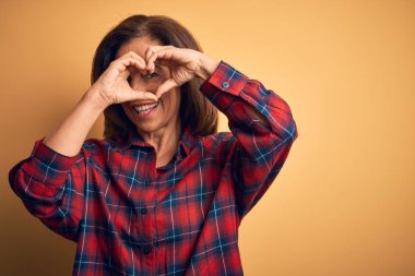Orta yaşlı, güzel bir kadın, gündelik gömleğini giymiş, izole edilmiş sarı arka plan üzerinde duruyor, el ve parmaklarıyla kalp şeklinde, tabelanın içinden bakıp gülümsüyor.