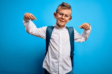 Zeki gözlüklü, mavi arka planda okul çantası takan, kendine güvenen, kendini gururlu ve mutlu gösteren küçük beyaz çocuk..
