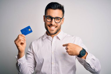 Young business man holding credit card over isolated background with surprise face pointing finger to himself