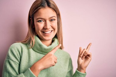 Sarışın, güzel bir kadın pembe yünlü bir arka planda kazak giyiyor gülümsüyor ve kameraya bakıyor. İki eliyle ve parmaklarıyla işaret ediyor..