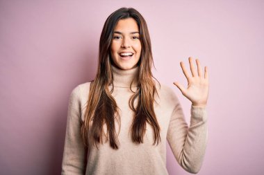 Balıkçı yaka kazak giyen genç ve güzel bir kız soyutlanmış pembe arka plan üzerinde duruyor ve parmaklarıyla beş numarayı gösterirken kendinden emin ve mutlu bir şekilde gülümsüyor..
