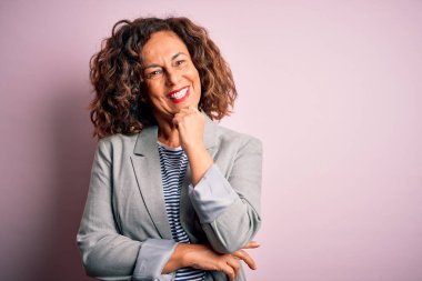 Soyutlanmış pembe arka planda zarif bir ceket giyen orta yaşlı güzel bir iş kadını kameraya kollarını kavuşturup gülümseyerek ve çenesini kaldırarak bakıyor. Olumlu düşünüyorum..